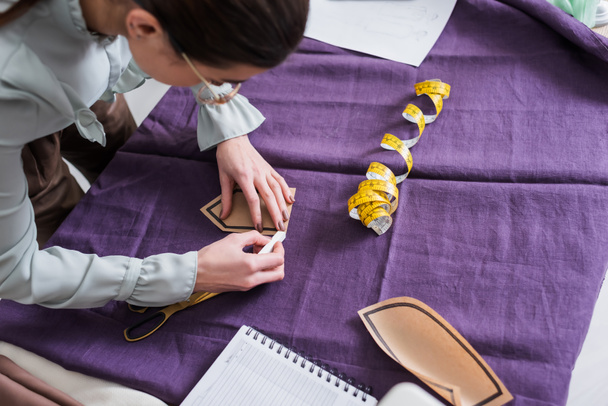 Overhead view of seamstress marking cloth and holding sewing pattern near scissors  - Photo, Image