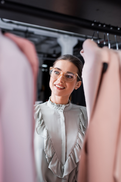 Smiling seamstress looking at blurred clothes on hangers  - Foto, Imagem