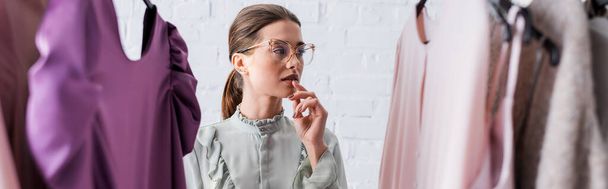 Pensive seamstress in eyeglasses standing near blurred clothes, banner  - Φωτογραφία, εικόνα