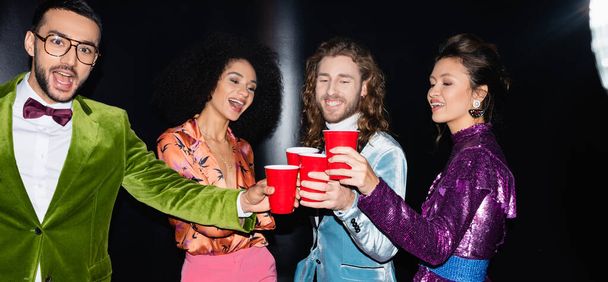 positive multirassische Freunde in bunten Klamotten, die aus Plastikbechern im Nachtclub auf schwarzem Hintergrund trinken, Banner - Foto, Bild