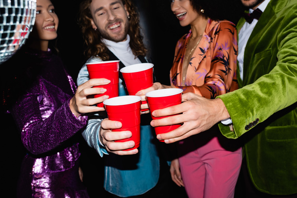positive multiracial friends in colorful clothes toasting with plastic cups in night club on black background - Фото, изображение