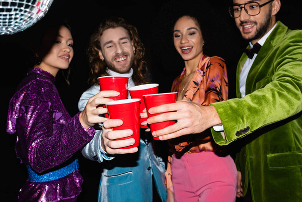smiling multiracial friends in colorful clothes toasting with plastic cups in night club on black background - Photo, image