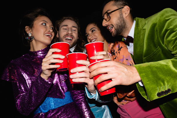 positive multiracial friends in colorful clothes toasting with plastic cups on black background - Fotografie, Obrázek