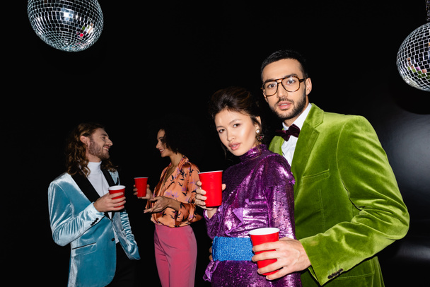 pozitivní multiraciální přátelé v barevných šatech stojící s plastovými poháry v rukou na černém pozadí - Fotografie, Obrázek