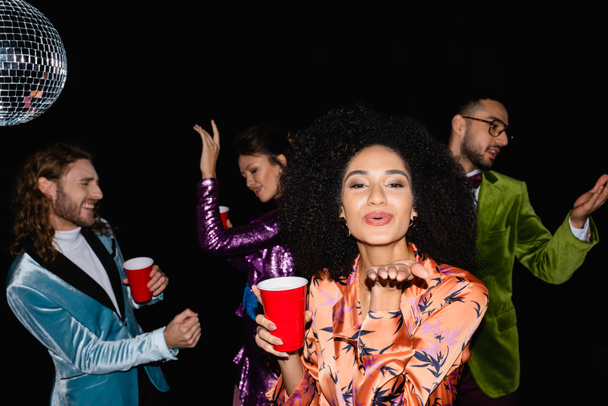 positivo afroamericano donna con soffiante bacio gesto sulla festa con amici multirazziali su sfondo nero - Foto, immagini