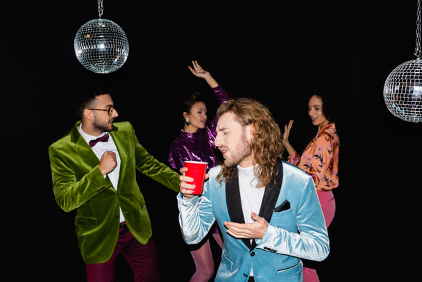 speelse jonge volwassen man met plastic beker en grimmig op partij met multiraciale vrienden dansen op zwarte achtergrond  - Foto, afbeelding
