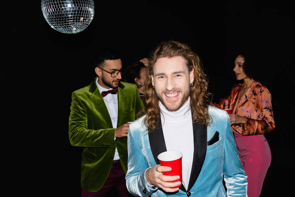 smiling young adult man holding plastic cup near blurred multiracial friends standing on black background - 写真・画像