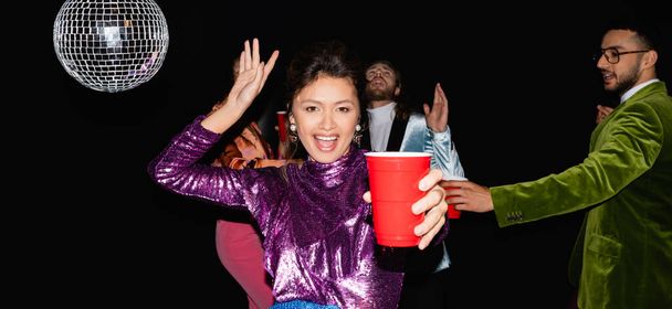 mujer asiática con taza de plástico bailando cerca borrosa multirracial amigos en ropa de colores sobre fondo negro, bandera - Foto, imagen