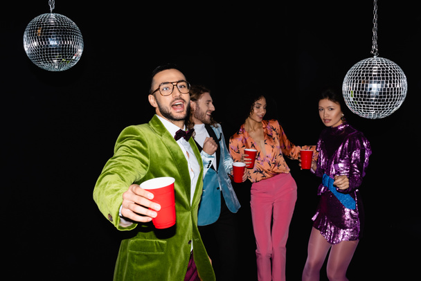 positiivinen arabi mies lasit tanssia monirotuisten ystävien kanssa värikkäissä vaatteissa yökerhossa mustalla taustalla - Valokuva, kuva