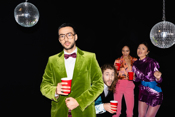 young adult arab man standing near funny multiracial friends in colorful clothes at party on black background - Foto, Imagem