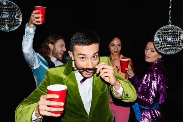 joven adulto árabe hombre ajuste gafas cerca divertido multiracial amigos en colorido ropa en la fiesta en negro fondo - Foto, Imagen