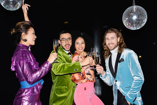 amigos multirraciales felices bailando con copas de champán sobre fondo negro - Foto, Imagen