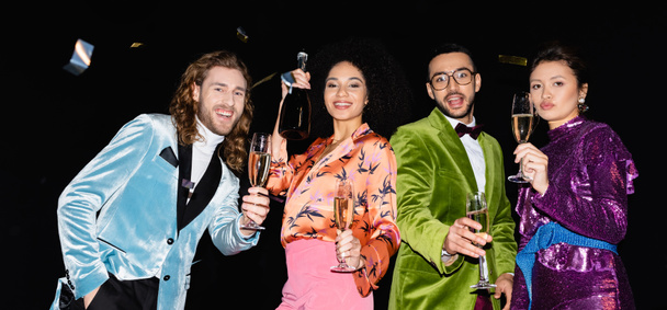 verspielte multirassische Freunde, die mit Champagnergläsern auf schwarzem Hintergrund tanzen, Banner - Foto, Bild