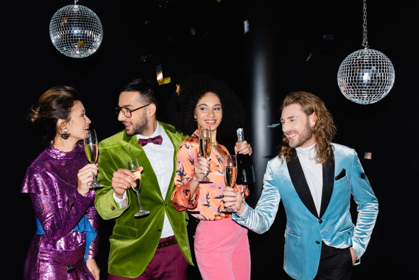 happy multiracial friends in colorful clothes with glasses of champagne having party on black background - Photo, Image
