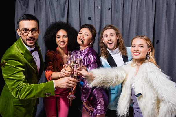 interracial friends in stylish clothes clinking with glasses of champagne near grey curtain on black background - Photo, Image