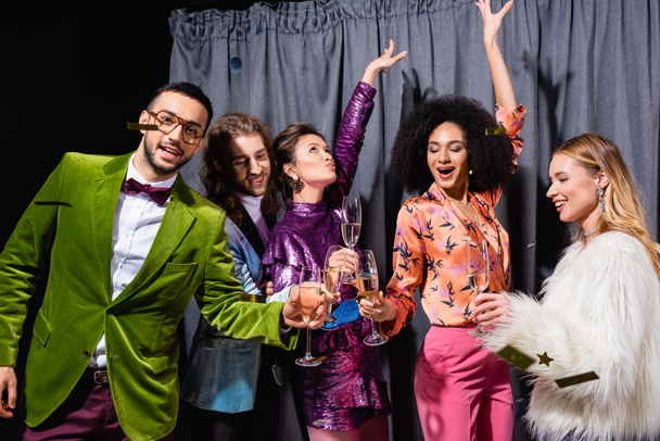 interracial přátelé ve stylovém oblečení pití šampaňského a tanec v blízkosti šedé opony na černém pozadí - Fotografie, Obrázek