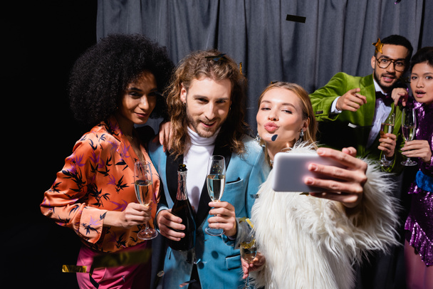 young adult interracial friends in bright clothes taking selfie on smartphone near grey curtain on black background - Photo, Image