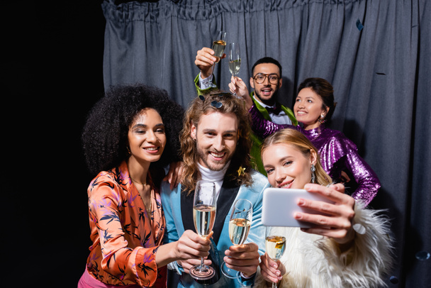 sonriente interracial amigos en brillante ropa tomando selfie en smartphone cerca gris cortina en negro fondo - Foto, Imagen