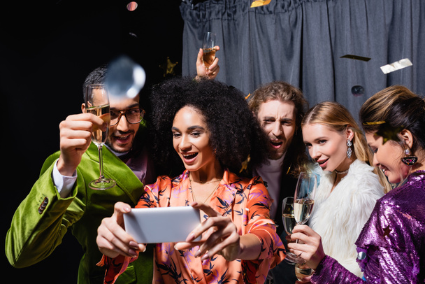 positive interrassische Freunde in heller Kleidung machen Selfie auf dem Smartphone in der Nähe des grauen Vorhangs auf schwarzem Hintergrund - Foto, Bild