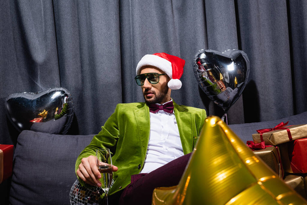 young adult arab man sitting on couch in santa hat and holding glass of champagne on grey background - Fotoğraf, Görsel