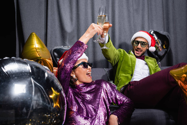 young adult arab man and asian woman sitting on couch with glasses of champagne near grey curtain on black background - Fotografie, Obrázek