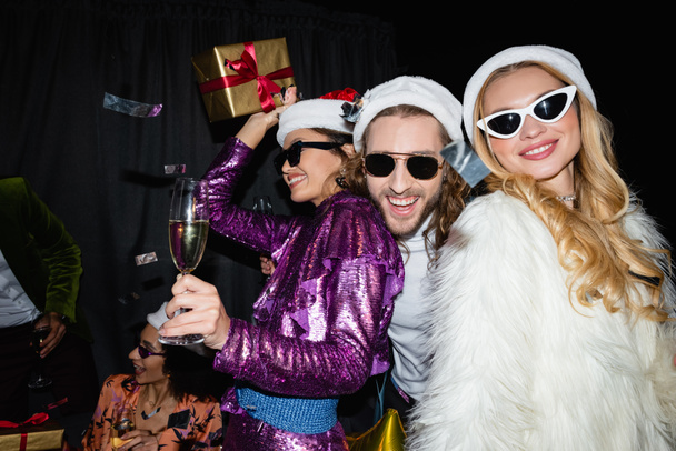 Ευτυχισμένοι διαφυλετικοί φίλοι με καπέλα santa πίνοντας σαμπάνια και γιορτάζοντας το νέο έτος σε γκρι φόντο - Φωτογραφία, εικόνα