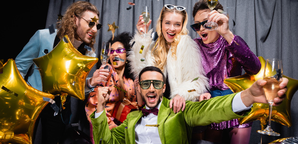 happy interracial friends in sunglasses and colorful clothes drinking champagne near grey curtain on black background, banner - Photo, Image