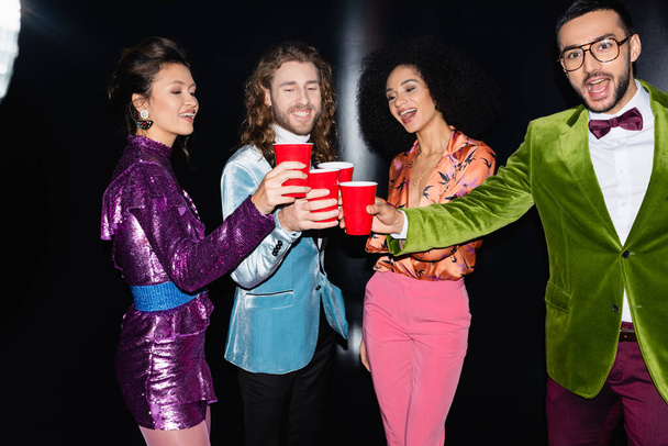 positive multiracial friends in colorful clothes drinking from plastic cups in night club on black background - Φωτογραφία, εικόνα