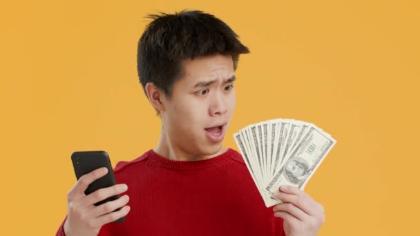 animado chinês cara usando smartphone segurando dinheiro no fundo amarelo - Filmagem, Vídeo