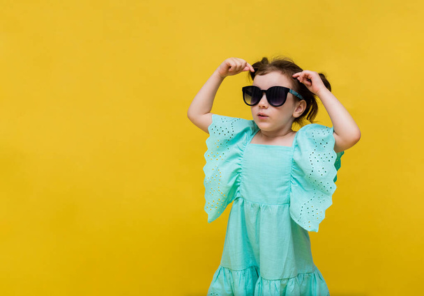 verrast klein meisje in een blauwe jurk en zonnebril op een gele achtergrond met ruimte voor tekst - Foto, afbeelding