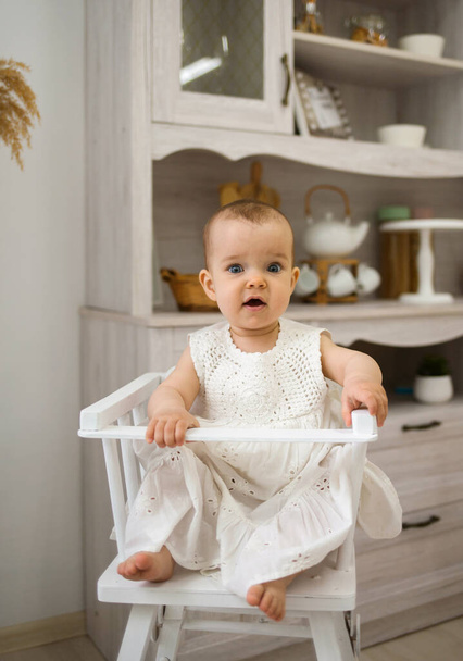 niña en una silla de bebé blanca se sienta en la cocina y mira a la cámara - Foto, imagen