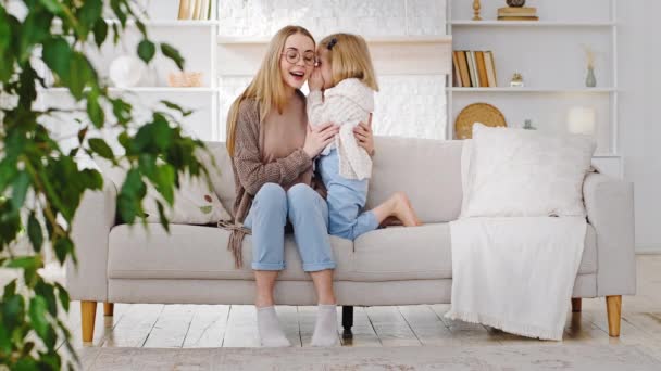 Young woman single mother and little cute daughter child sitting on sofa at home, baby whispers secret in ear talks tells secrecy information dream gossip, family chatting casual conversation indoor - Footage, Video