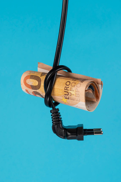 Een biljet van vijftig euro wordt in een knoop van een stroomkabel met stekker gestoken. Een close-up. Blauwe achtergrond. Elektro-efficiëntie en energiebesparing - Foto, afbeelding
