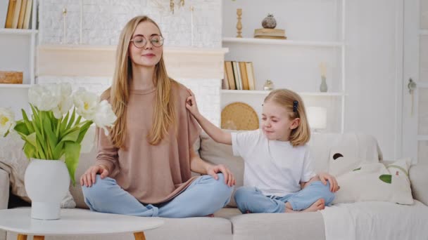 Valkoihoinen perhe nuori rauhallinen äiti nainen blondi meditoi taapero tyttö lapsi tytär istua lootus asennossa sohvalla pieni lapsi tekee melua työntää äiti häiritsee keskittymistä huono käyttäytyminen - Materiaali, video