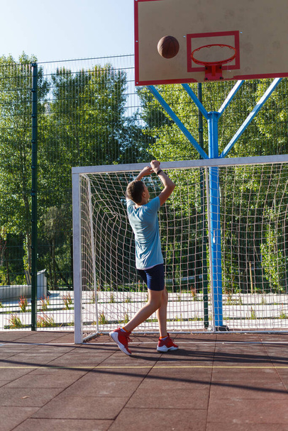 Симпатичный мальчик в зеленой футболке играет в баскетбол на городской площадке. Активный подросток наслаждается игрой на открытом воздухе с оранжевым мячом. Хобби, активный образ жизни, спорт для детей. - Фото, изображение