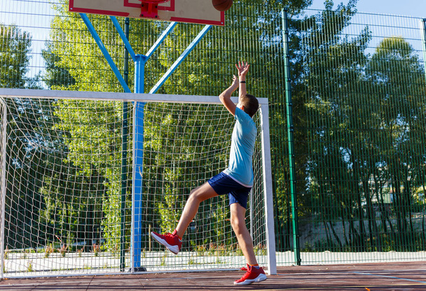 Ein süßer Junge im grünen T-Shirt spielt Basketball auf einem städtischen Spielplatz. Aktive Teenager genießen ein Spiel im Freien mit einem orangefarbenen Ball. Hobby, aktiver Lebensstil, Sport für Kinder. - Foto, Bild