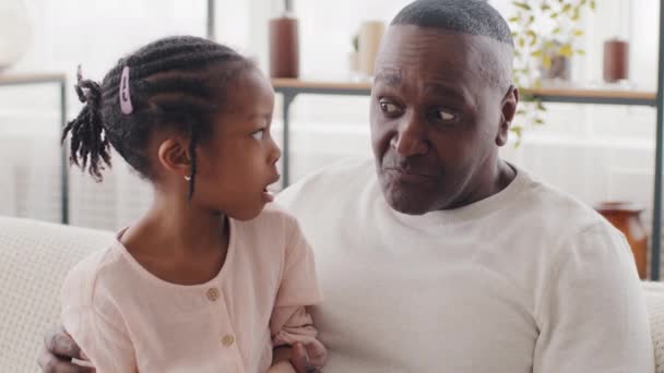 Портрет смешанной расы семьи два человека афро маленькая девочка дочь школьница с черным этническим взрослым взрослым мужчиной пожилой человек отец дедушка разговаривает болтая разговор сидя на диване дома - Кадры, видео