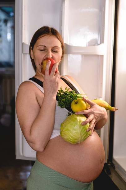 Pregnant woman holding fresh fruits near the refrigerator. Woman eating a red apple. - Foto, afbeelding