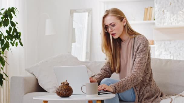 Молода тисячолітня біла дівчина блондинка фрілансер студент друкує звіт на ноутбуці, що сидить низько батареї при комп'ютерних з'єднаннях проблеми втрачають відчуття шокової невдачі, покриваючи рот руками вдома
 - Кадри, відео