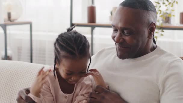 Glücklich afrikanisch-amerikanische Familie reifer Mann mit Falten Vater Großvater mit kleiner Tochter Mädchen Kind sitzt zusammen zu Hause auf der Couch reden lachen kommunizierende Gespräch Händchen haltend - Filmmaterial, Video
