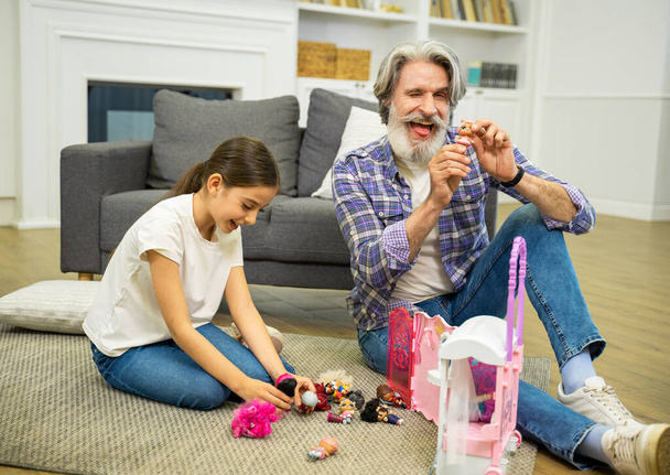 Улыбающийся дедушка веселится и играет с внучкой, показывая ему кукольный домик - Фото, изображение