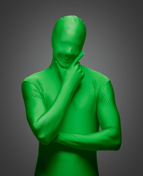 Hombre pensante con la mano en la barbilla con traje de nylon verde completo sobre un fondo gris - Foto, Imagen