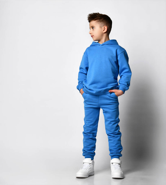 Full-length portrait of a young handsome guy with a stylish haircut in pastel modern blue tones, with a hood, a sweatshirt, standing, hands in pockets - Fotografie, Obrázek