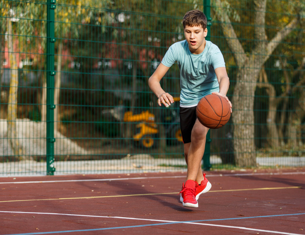A cute boy in a green t-shirt plays basketball on a city playground. Active teen enjoying an outdoor game with an orange ball. Hobby, active lifestyle, sport for kids. - Zdjęcie, obraz