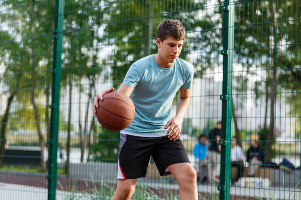 Un chico lindo con una camiseta verde juega baloncesto en un parque de la ciudad. Adolescente activo disfrutando de un juego al aire libre con una bola naranja. Aficion, estilo de vida activo, deporte para niños. - Foto, Imagen