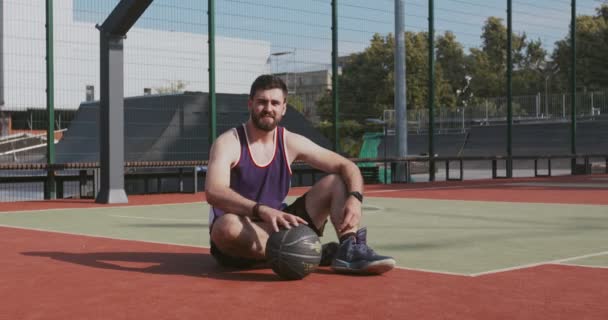 Открытый портрет молодого активного баскетболиста, сидящего на полу корта и играющего с мячом, смотрящего в камеру - Кадры, видео