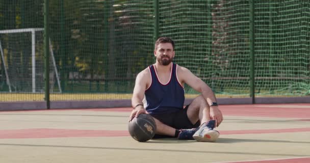 Jonge Kaukasische streetball speler zittend op speelplaats vloer en spelen met bal, kijken naar camera, slow motion - Video