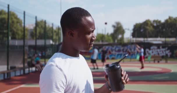 Молодой африканский американец пьет воду из спортивной бутылки на открытой баскетбольной площадке, вид сбоку, замедленная съемка - Кадры, видео