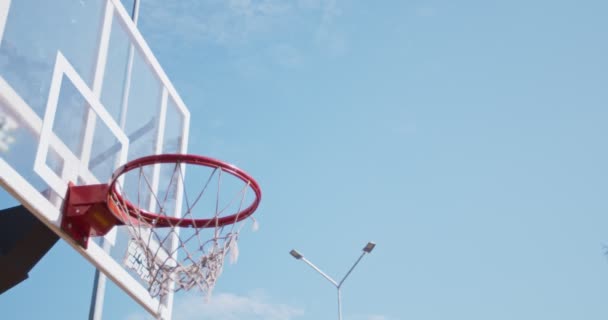 Στόχος και επιτυχία. Εξωτερική βολή της μαύρης μπάλας του μπάσκετ που πέφτει σε streetball βρόχο πάνω από το μπλε φόντο του ουρανού - Πλάνα, βίντεο