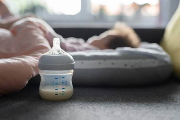 Enfoque selectivo de cerca en la leche materna o de fórmula en el biberón en la cama en casa con el bebé dormido en segundo plano: espacio para copiar el concepto de alimentación y lactancia para recién nacidos - Foto, imagen